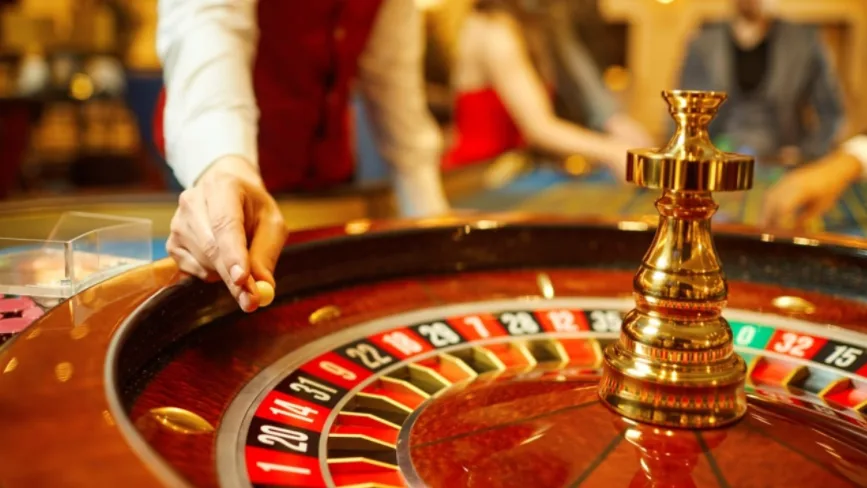 Cilindro de ruleta iluminado con la mano de una crupier a un costado y jugadores desenfocados al fondo.