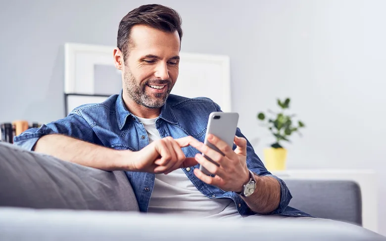 Fotografía del blog oficial de rojabet: un hombre revisa su teléfono sentado en un sofá.